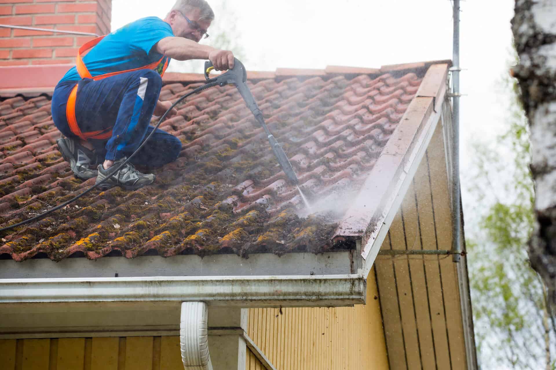 Caucasian man is washing the roof with a high pressure washer. He is wearing safety harness on a slippery roof.